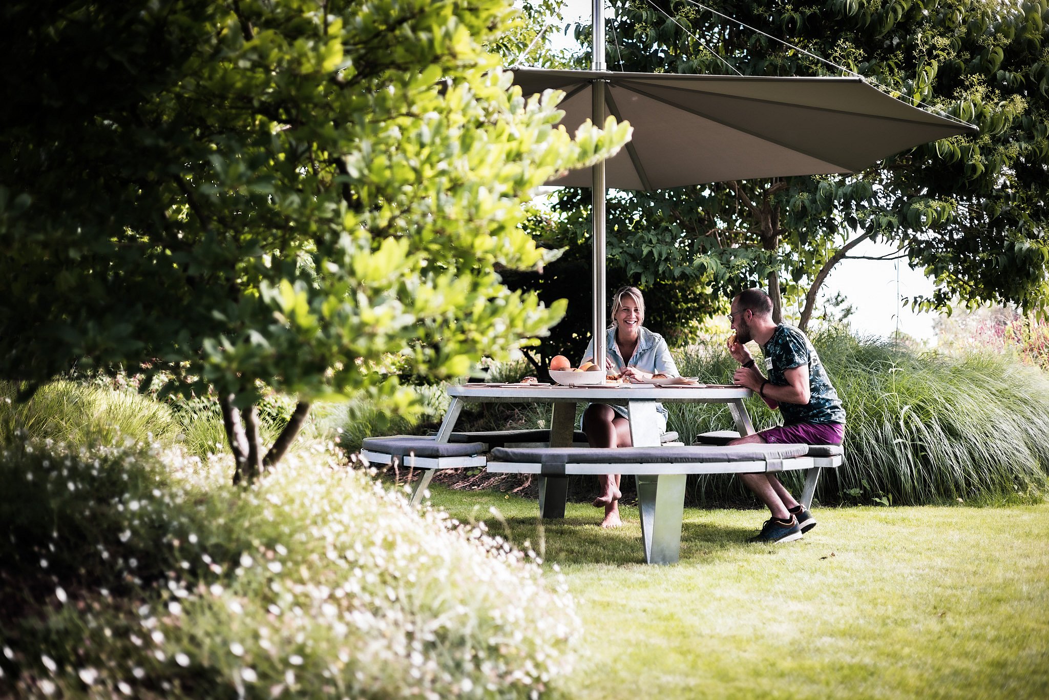 Een ronde Pantagruel picknicktafel met gebogen kussenbanken zit onder een witte Inumbra-tint in een weelderige tuin, omlijst door grassen en lommerrijke bomen