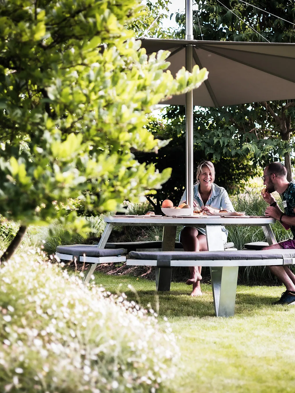 Een ronde Pantagruel picknicktafel met gebogen kussenbanken zit onder een witte Inumbra-tint in een weelderige tuin, omlijst door grassen en lommerrijke bomen