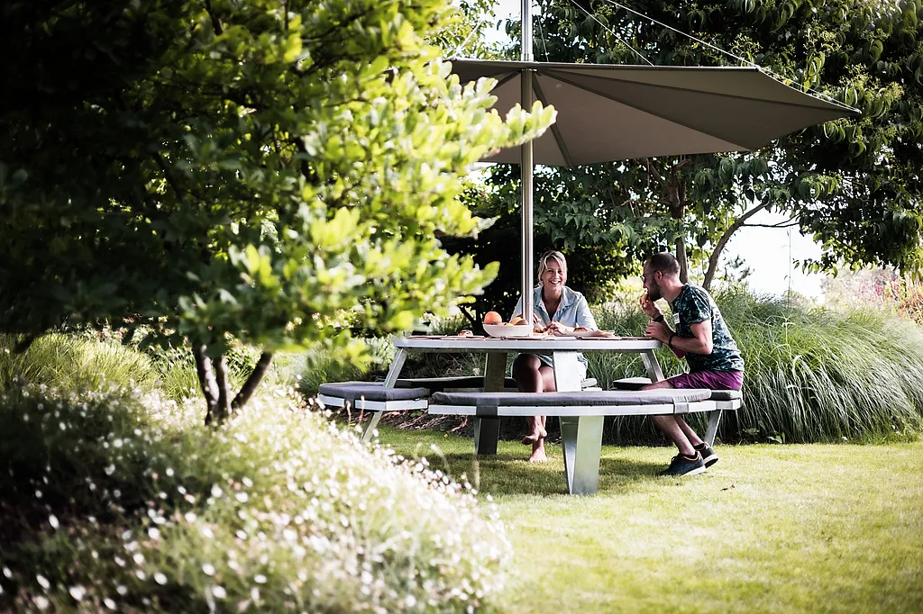 Ein runder Pantagruel-Picknicktisch mit geschwungenen gepolsterten Bänken steht unter einem weißen Inumbra-Schatten in einem üppigen Garten, eingerahmt von Gräsern und grünen Bäumen