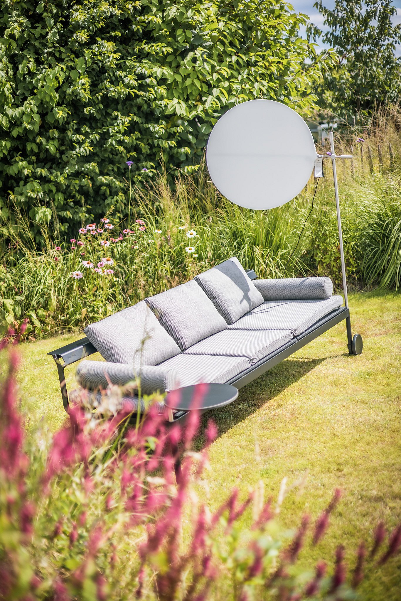 A grey Sol+Luna sofa with a round side table and circular lamp stands among tall grasses and flowering borders in the garden.