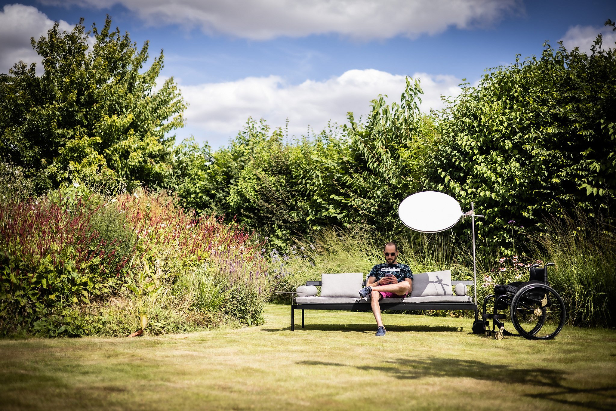 A man sits reading on a grey Sol+Luna sofa in the garden, shaded by the circular lamp.