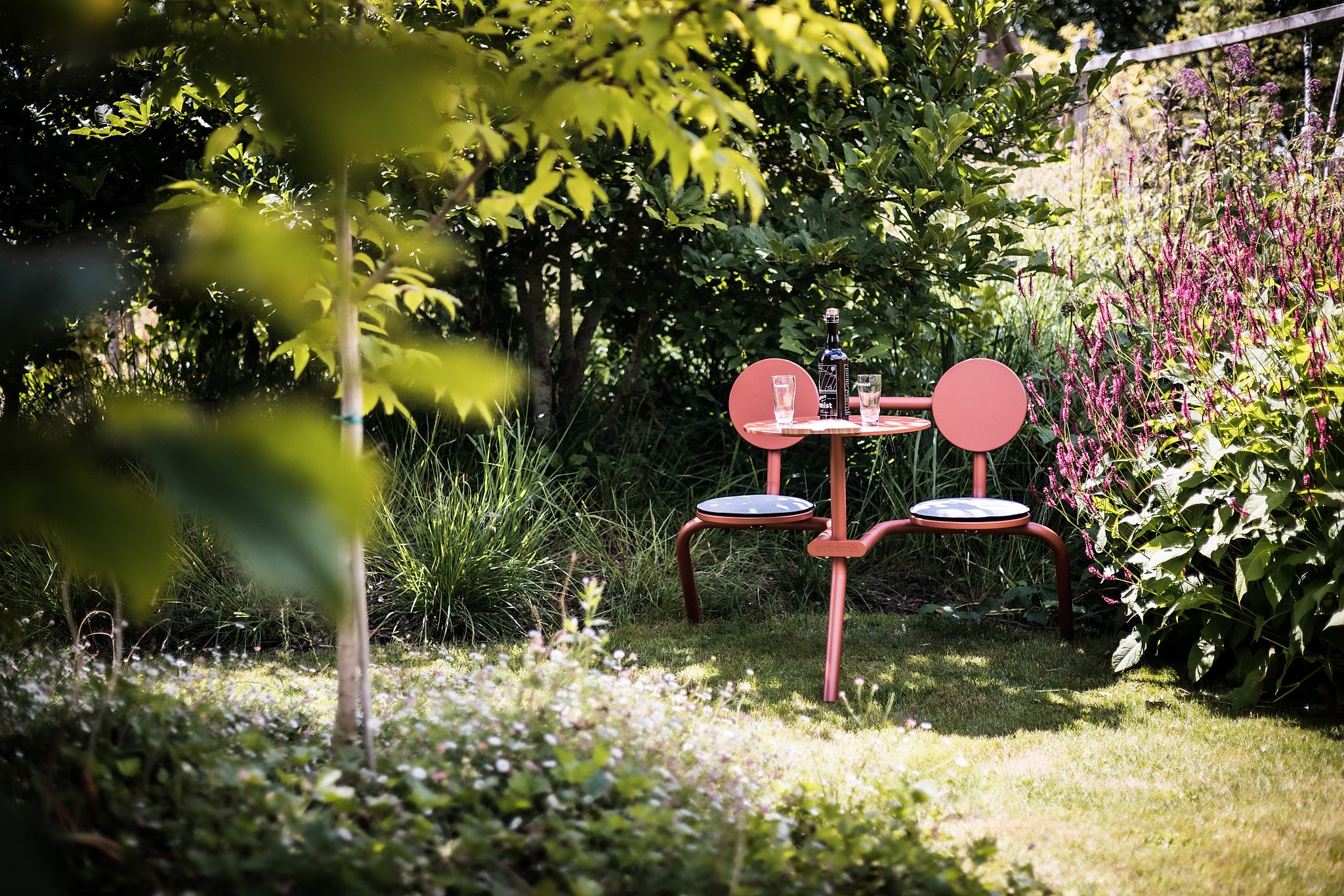 A copper-brown Bistroo table with two integrated seats is tucked into a planted garden corner, set with a bottle and two glasses.