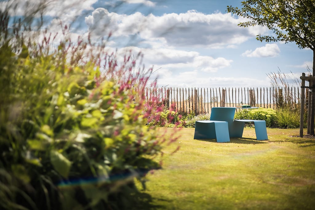 A sky-blue Picnik seat-and-table unit sits on a lawn beyond flowering borders, with a rustic fence and open sky in the background