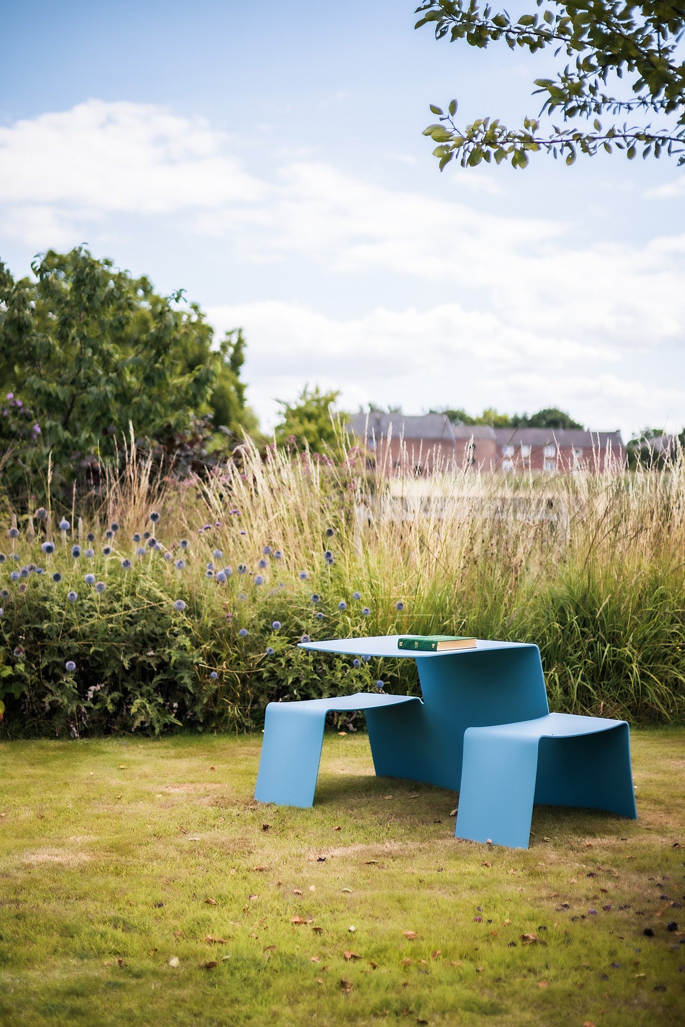 Close view of a sky-blue Picnik unit on the grass, with a book resting on the tabletop and tall meadow planting behind.
