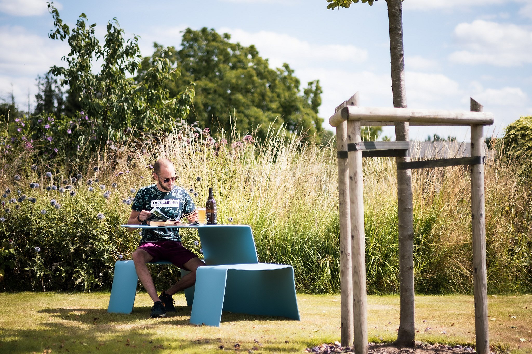 A man reads at a sky-blue Picnik unit on the lawn, surrounded by tall ornamental grasses and summer planting.