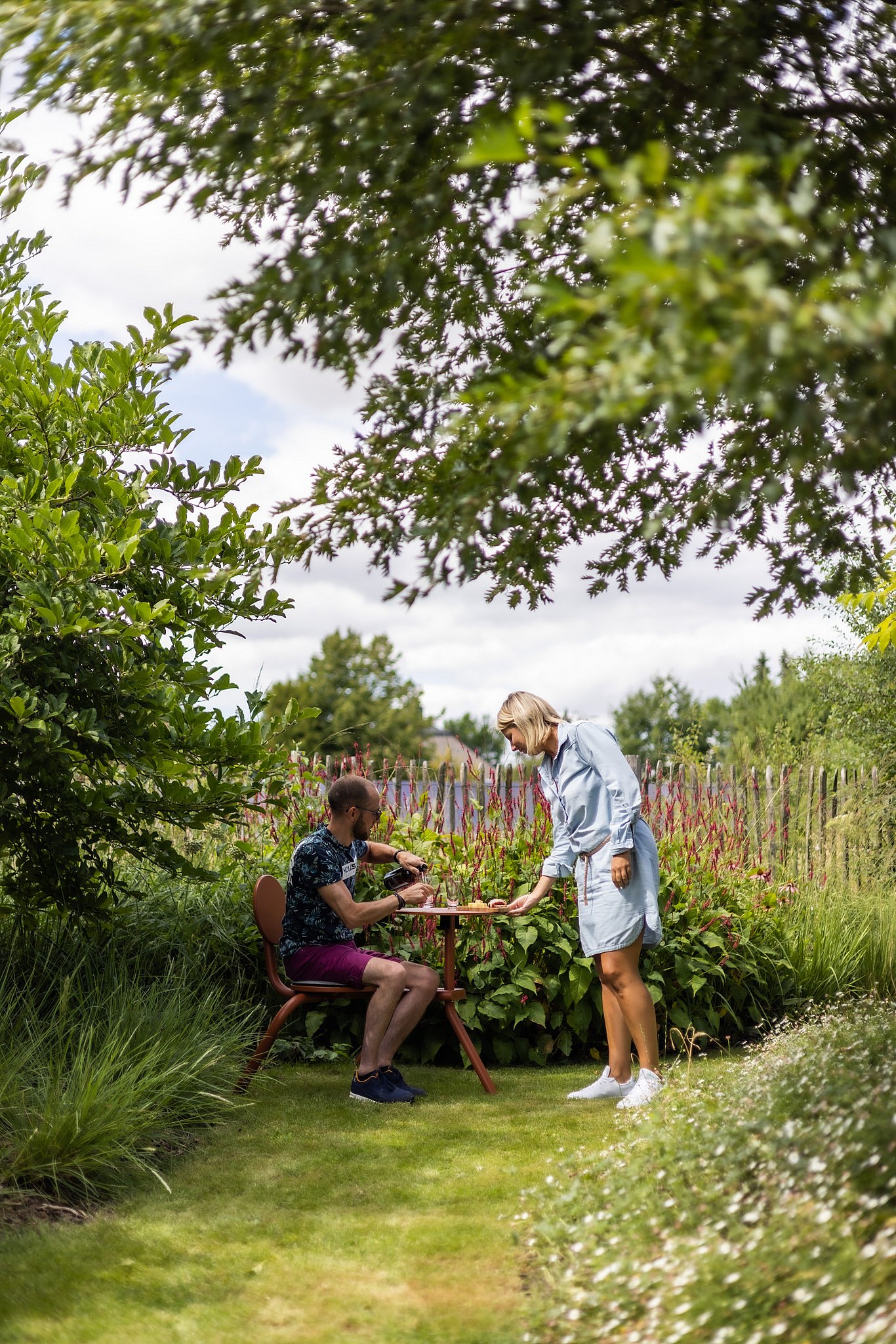 A woman serves a seated man at a copper-brown Bistroo table beneath overhanging branches and flowering borders.