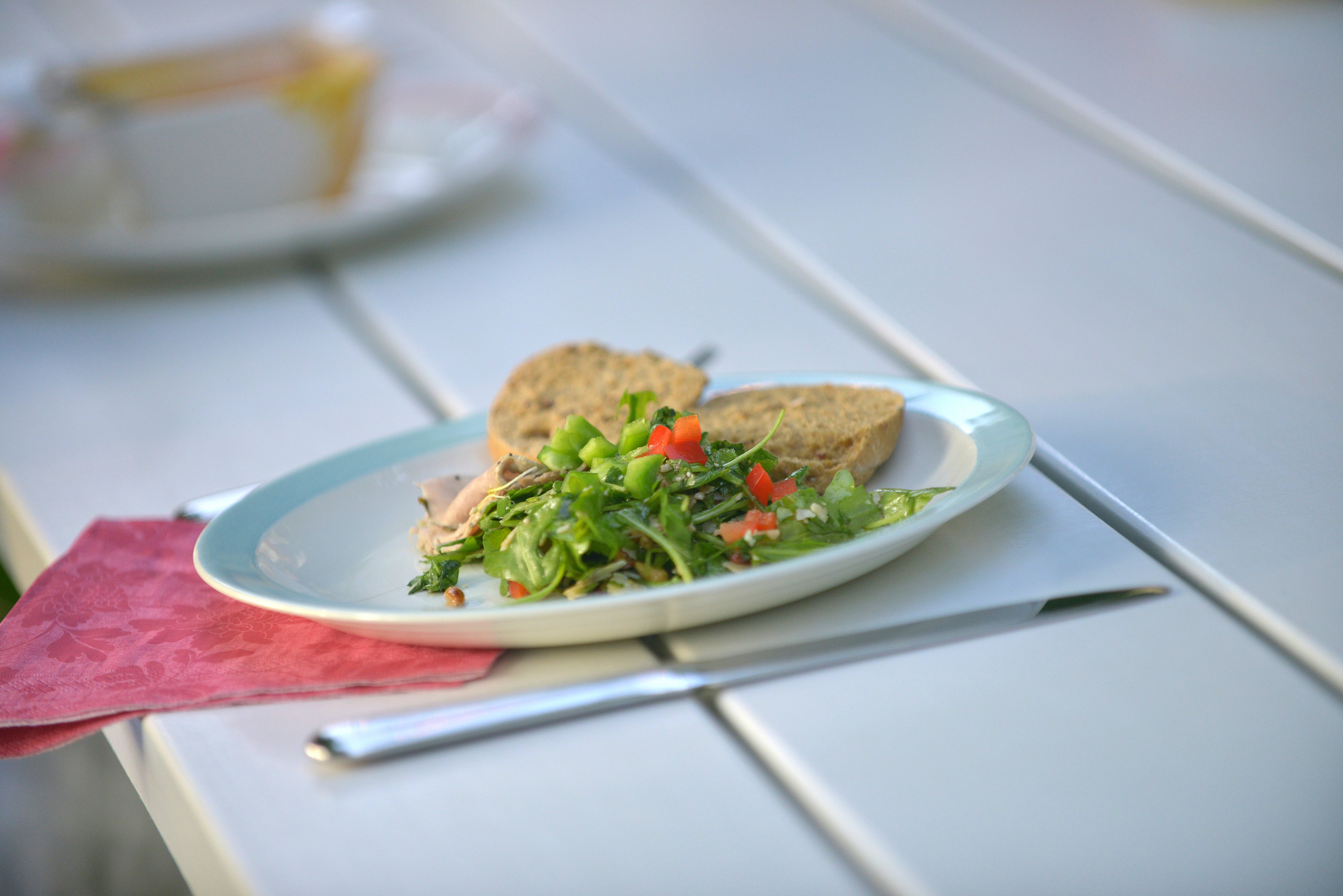 Nahaufnahme eines Tellers mit Salat und Brotscheiben, die auf der weißen Tischplatte neben Besteck und einer rosa Serviette ruhen.