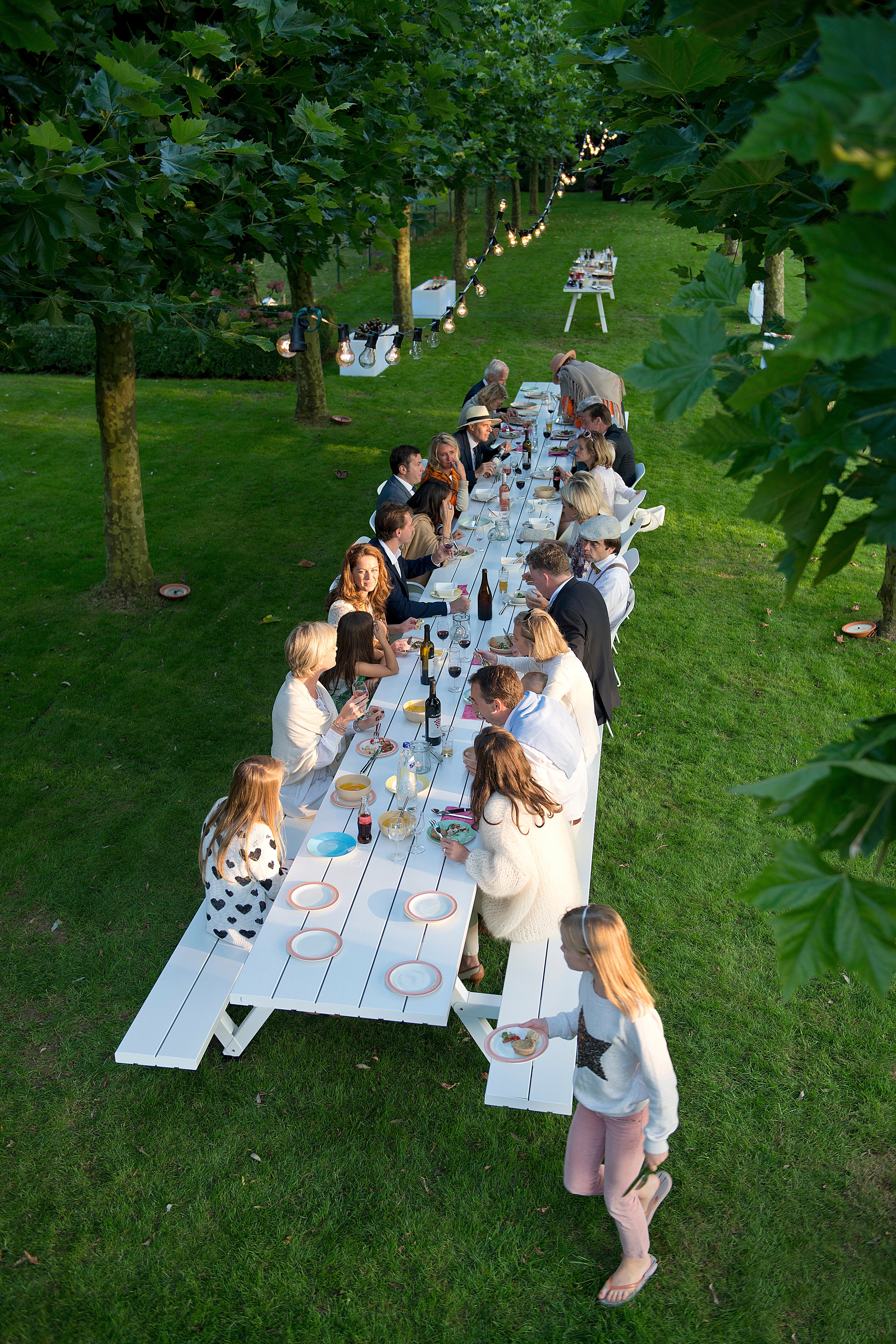 Luchtfoto van een extra lange witte Marina picknicktafel gedekt voor een tuinfeest bij schemering, met lichtsnoeren in de bomen en een grote groep volwassenen en kinderen die samen eten.