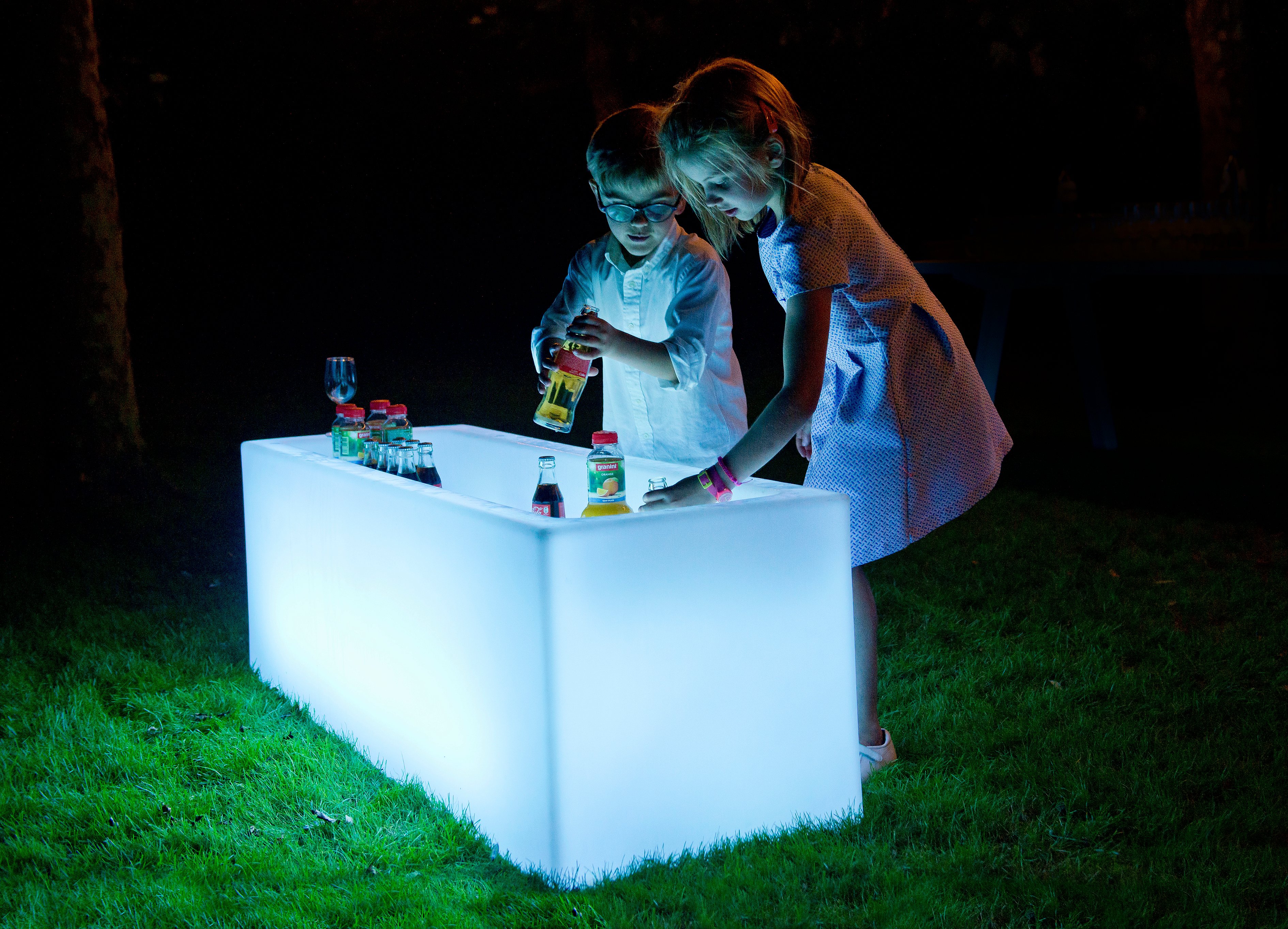 Zwei Kinder wählen Getränke aus einer strahlend weißen Icecube-Kühlbox aus, die nachts auf dem Rasen steht.