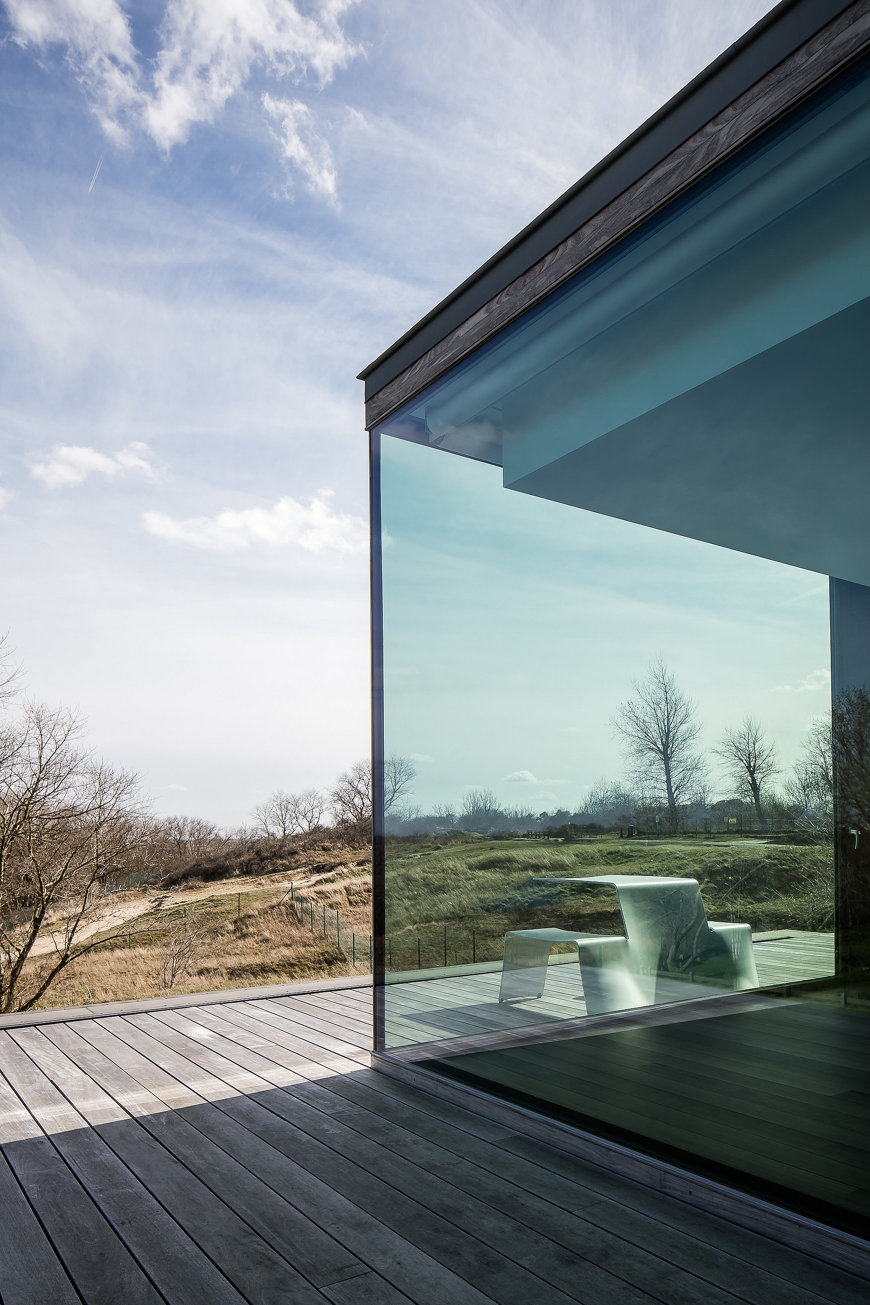 In der Ecke einer Glasfassade spiegelt sich ein weißer Picnik-Tisch, neben dem Haus erstrecken sich die Dünenlandschaft und die Holzterrasse.