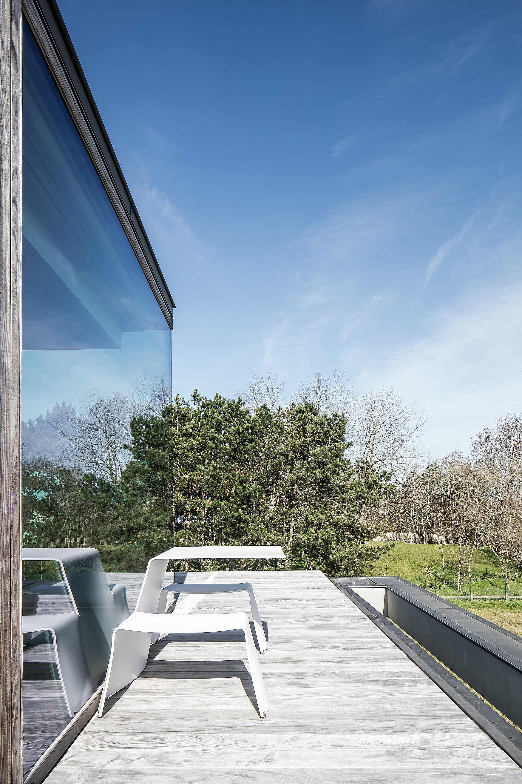 Ein weißer Picnik-Tisch mit zwei integrierten Sitzen steht auf einer verwitterten Holzterrasse neben einem Schlafzimmer mit Glaswänden und bietet Blick auf Dünenvegetation und Bäume.