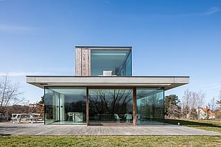 Weitblick auf ein modernes, niedriges Haus mit raumhoher Verglasung und breitem Holzdeck, mit einem weißen Gargantua-Picknicktisch auf der Terrasse links.