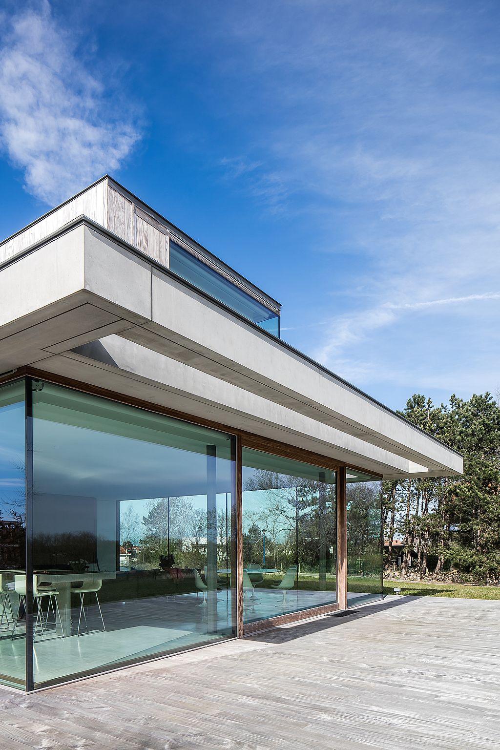 Seitenansicht des modernen Hauses mit tiefen Dachüberhängen und Vollverglasung, das sich zu einer breiten Holzterrasse mit minimalistischen weißen Möbeln innen und außen öffnet.