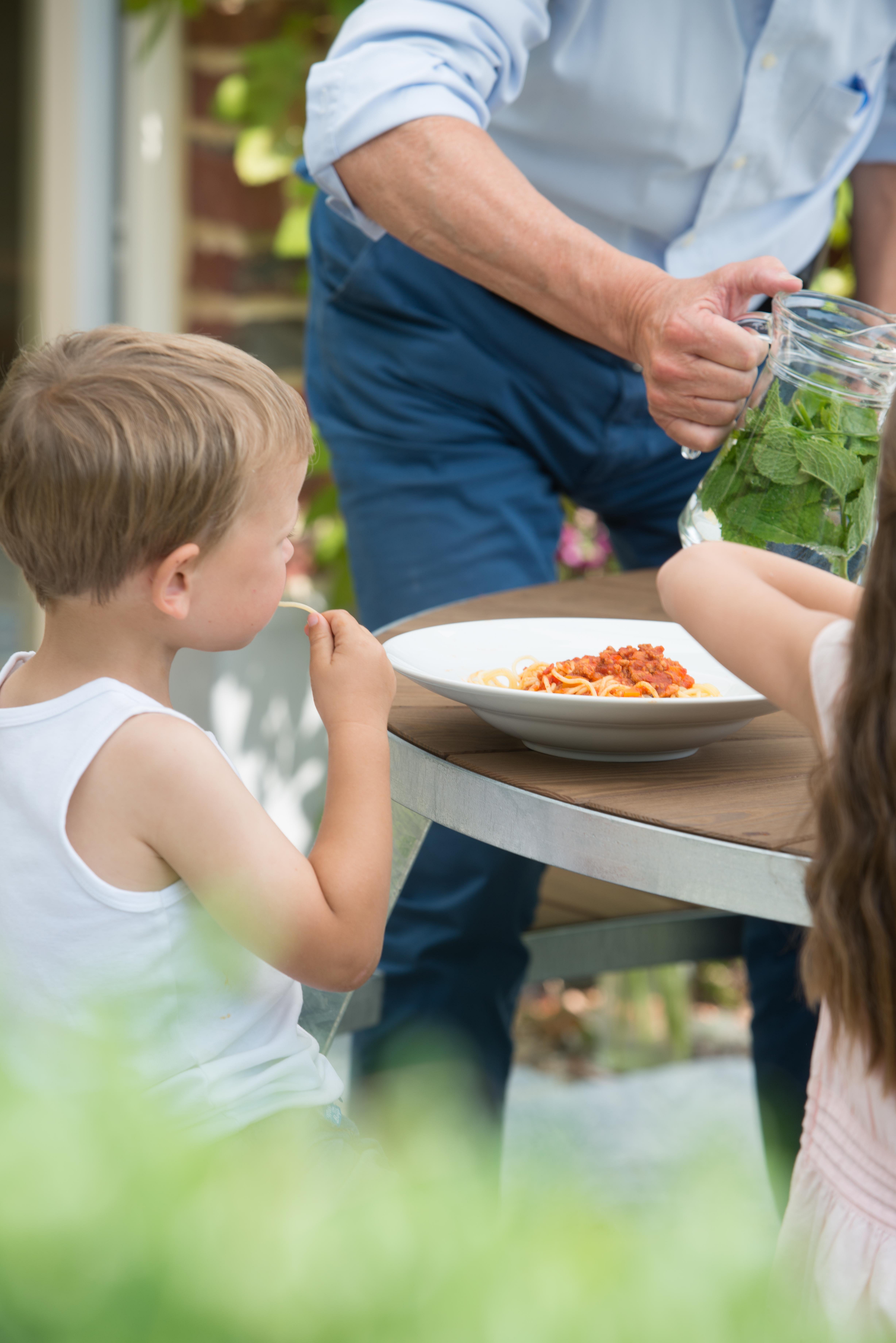 Nahaufnahme eines kleinen Jungen, der am Pantagruel-Tisch Spaghetti isst, während ein Erwachsener mit Kräutern angereichertes Wasser aus einem Glaskrug neben einen Teller Nudeln gießt.