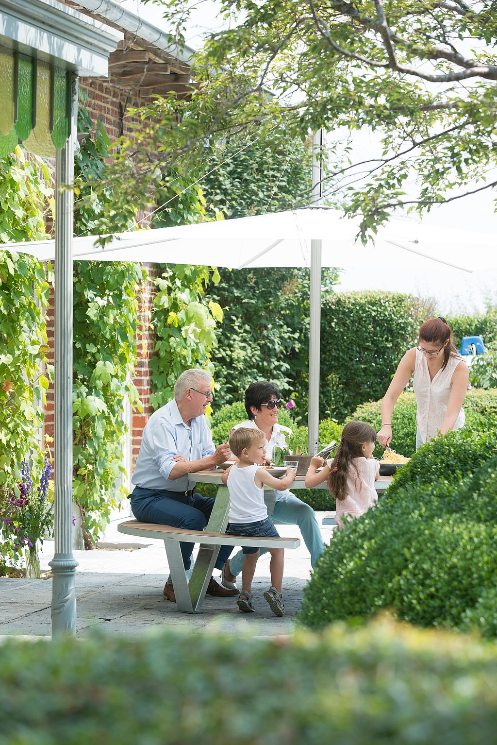 Seitenansicht einer Familie, die gemeinsam Pasta an einem Pantagruel-Picknicktisch unter einem weißen Inumbra-Sonnenschirm genießt, eingerahmt von Hecken und Gartenbepflanzungen.