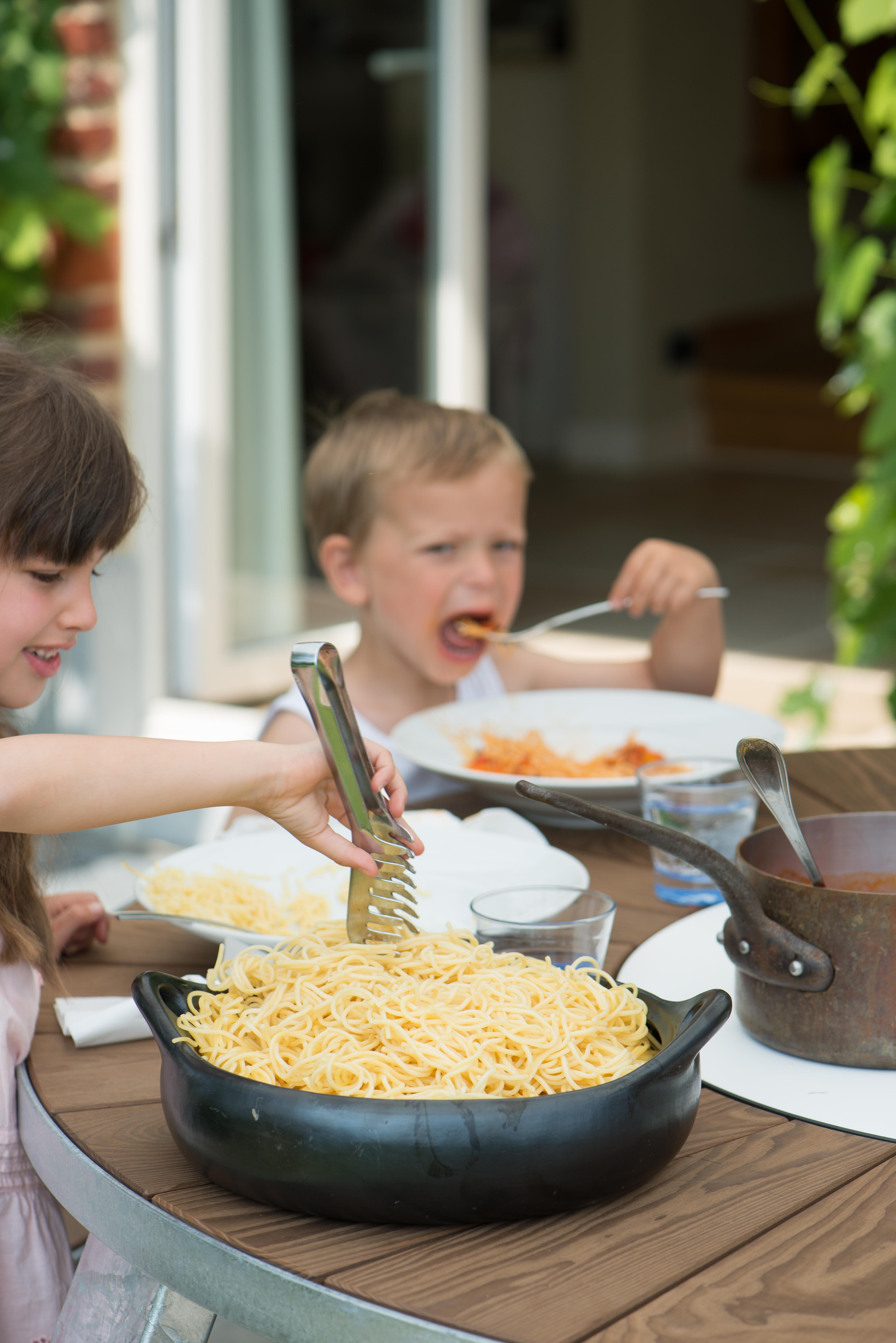 Nahaufnahme einer großen Pfanne Spaghetti auf der Pantagruel-Tischplatte, während zwei Kinder sich selbst bedienen und im Freien essen.