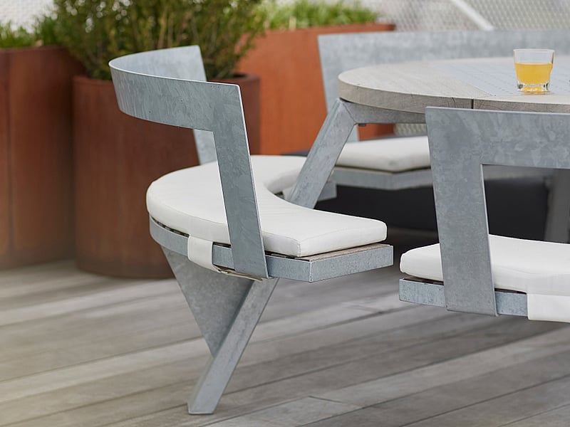 Close-up of a galvanized outdoor table and chair with pale seat cushions on a terrace, with large rust-colored planters and a drink on the tabletop.