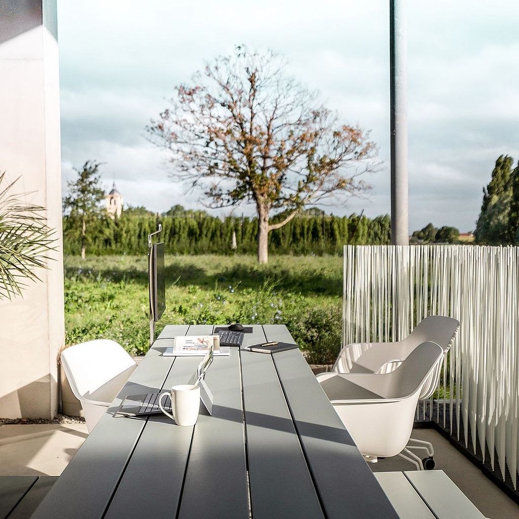 “A long pale grey outdoor worktable with integrated benches sits on a covered terrace, styled with laptops, a monitor, notebooks and coffee, overlooking a green field.