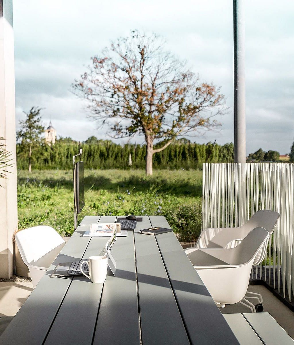 “A long pale grey outdoor worktable with integrated benches sits on a covered terrace, styled with laptops, a monitor, notebooks and coffee, overlooking a green field.