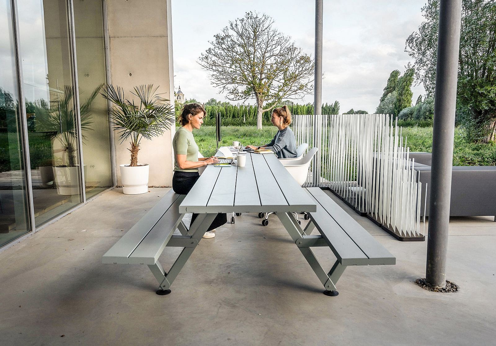   Moderner Outdoor-Arbeitsbereich mit einem langen, schattigen Tisch, weißen Stühlen, Lounge-Sitzgelegenheiten und einem Monitor, eingebettet zwischen hohen Pflanzkästen neben einem modernen Gebäude.
