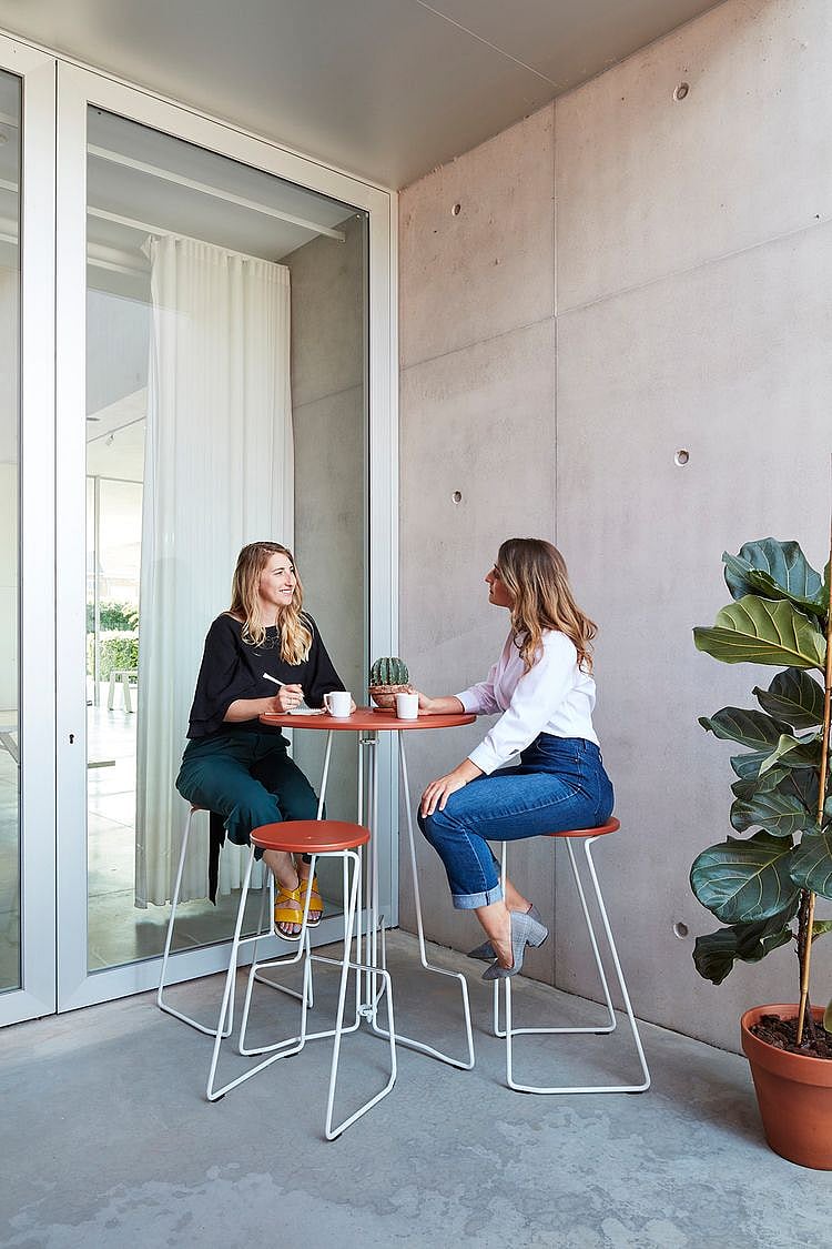 Two women chat over coffee at a terracotta-red high outdoor table with matching stools in a sheltered concrete corner beside a large potted plant.