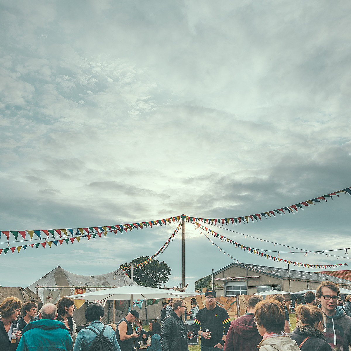 Crowd gathered at an outdoor festival beneath colorful bunting and string lights under a cloudy sky.