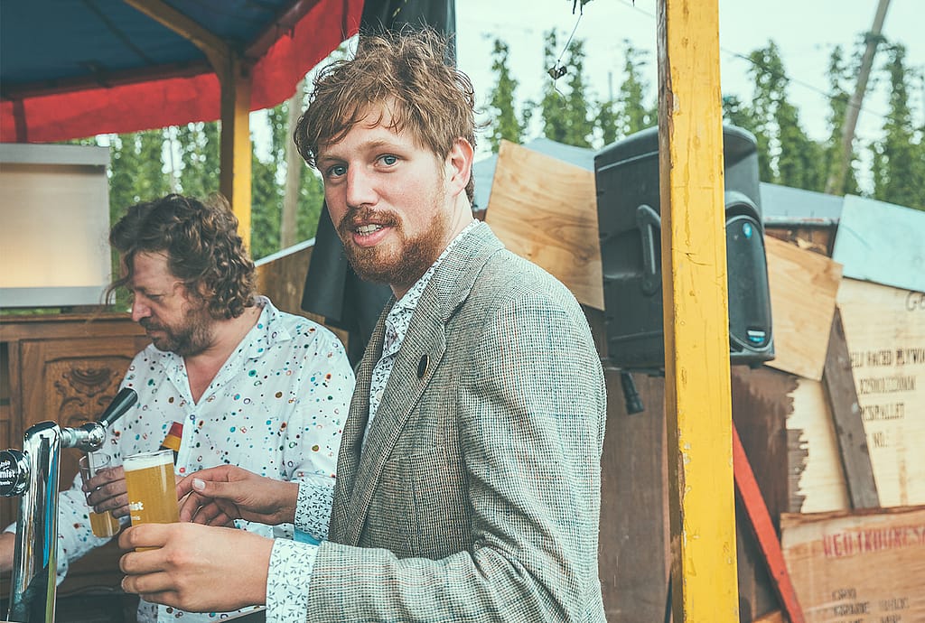 Thomas in einem Blazer hält ein Glas Bier neben dem Zapfhahn, hinter ihm gießt ein anderer Mann Bier ein.