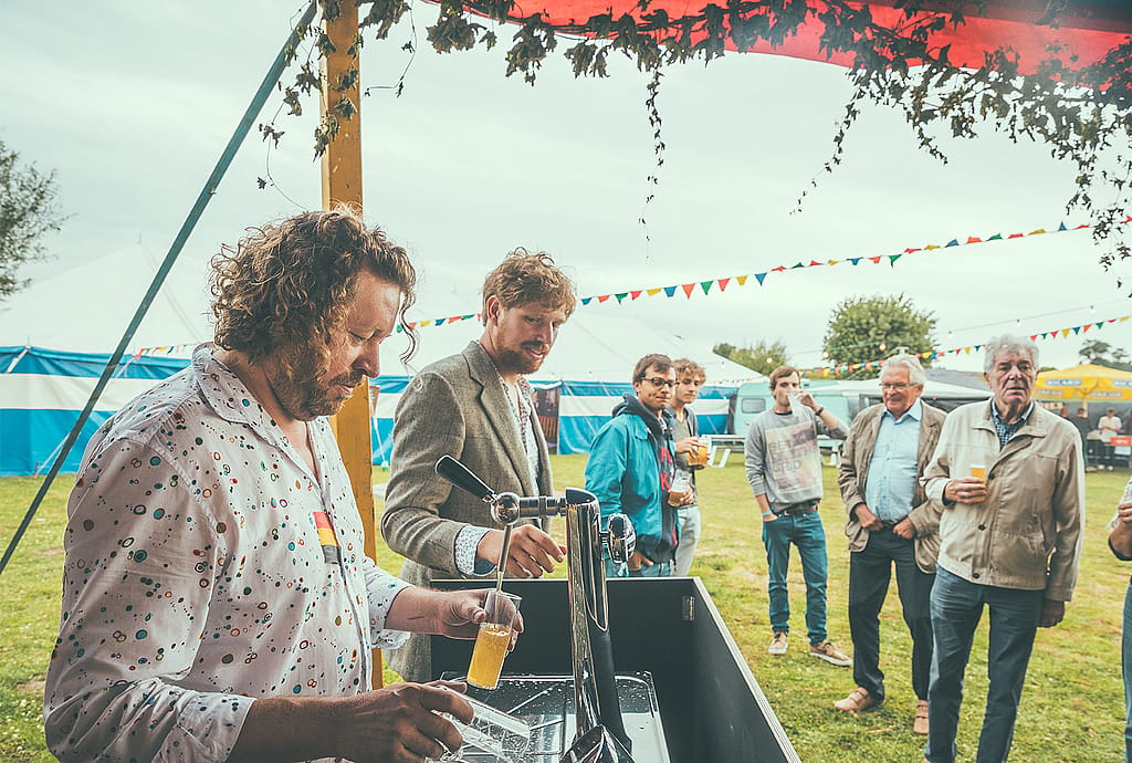 Dirk gießt Bier aus einem Fasshahn ein, während die Gäste in einer grasbewachsenen Festivalumgebung im Freien zusehen.
