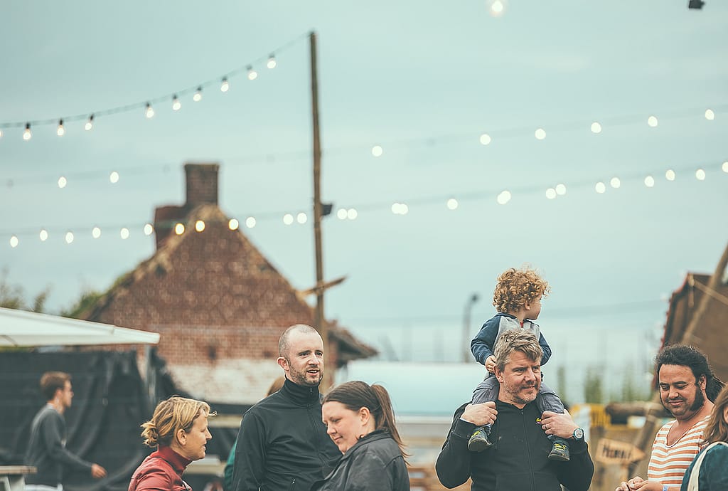 Festivalbesucher unterhalten sich im Freien unter Lichterketten, darunter ein Kind, das auf den Schultern eines Erwachsenen sitzt.