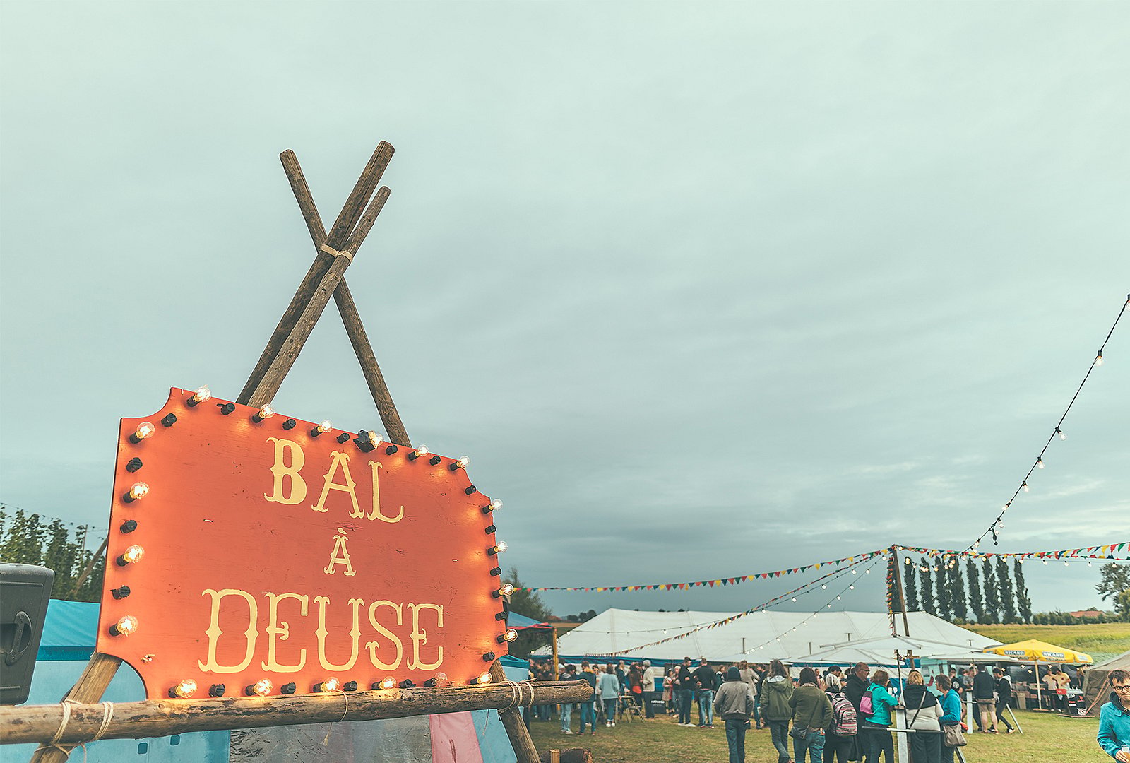 Beleuchtetes “Bal à Deuse” Festschild im Vordergrund mit Zelten, Wimpel und Besuchern dahinter.
