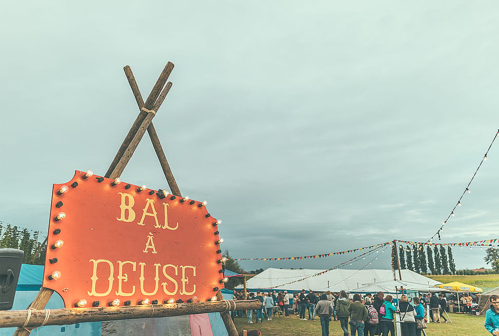 Beleuchtetes “Bal à Deuse” Festschild im Vordergrund mit Zelten, Wimpel und Besuchern dahinter.