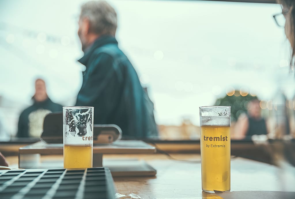 Zwei teilweise gefüllte Tremist-Biergläser auf einer Theke mit verschwommenen Festivalbesuchern im Hintergrund.