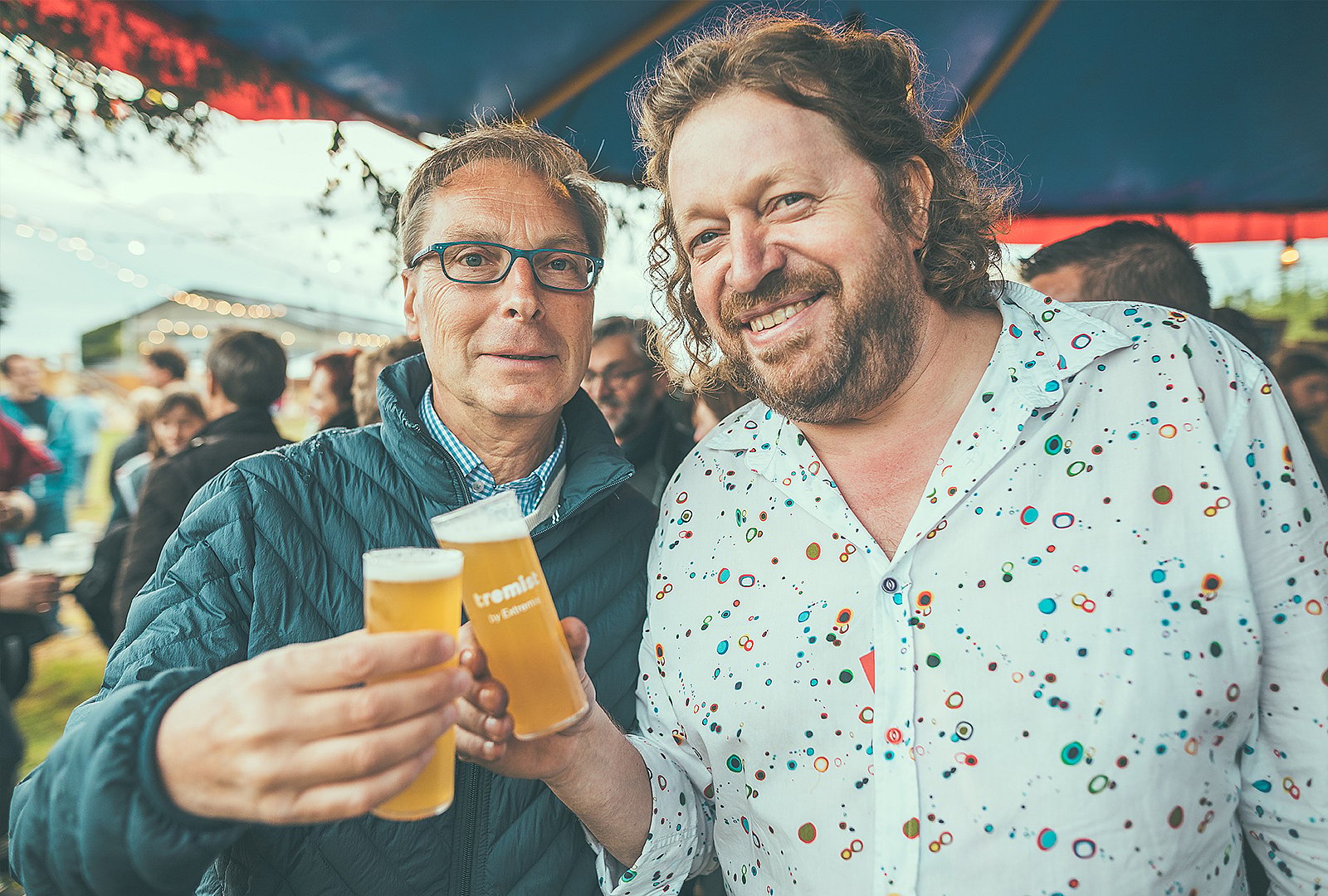 Zwei Männer lächeln und stoßen unter dem Festivalzelt auf Markenbiergläser in Richtung Kamera an.