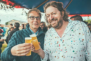 Zwei Männer lächeln und stoßen unter dem Festivalzelt auf Markenbiergläser in Richtung Kamera an.