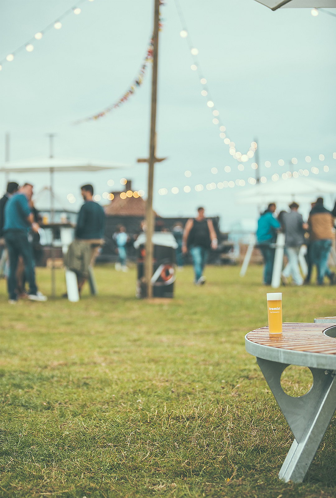 Einzelnes Glas Tremist-Bier am Rand eines runden Tisches im Freien, hinter dem Gäste und Lichter sanft verschwommen sind