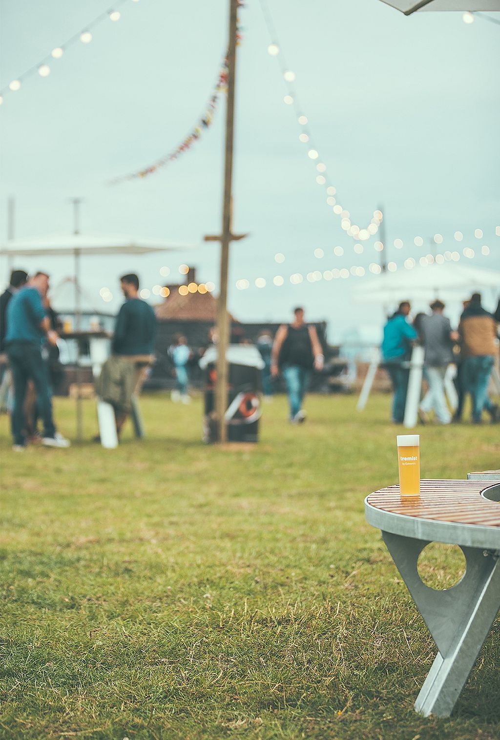 Einzelnes Glas Tremist-Bier am Rand eines runden Tisches im Freien, hinter dem Gäste und Lichter sanft verschwommen sind