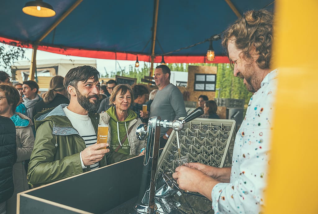 Gast hält ein Markenbierglas an der Bar, während Dirk hinter dem Zughahn unter dem Zelt steht.
