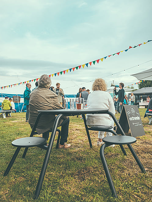 Zwei Personen sitzen mit Getränken an einem schwarzen Tisch im Freien und beobachten das Festival auf der anderen Seite des Feldes.