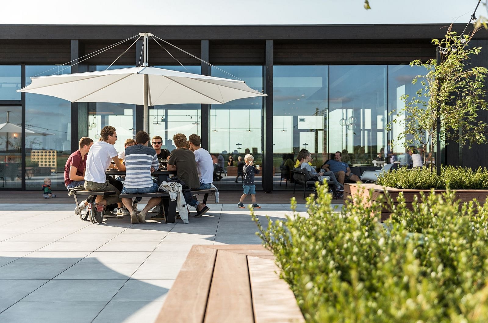 Grote witte inumbra parasols over ronde pantagruel picknicktafels op een terras, met mensen die buiten een modern gebouw zitten.