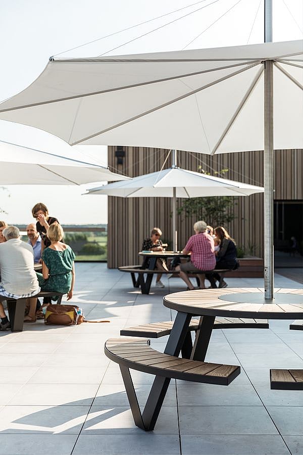 Grote witte inumbra parasols over ronde pantagruel picknicktafels op een terras, met mensen die buiten een modern gebouw zitten.