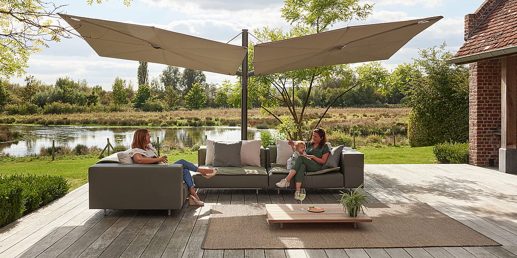 Twee volwassenen en een kind ontspannen op modulaire walrussen in de buitenlucht, zitten onder dubbele luifels op een terras met uitzicht op een vijver.