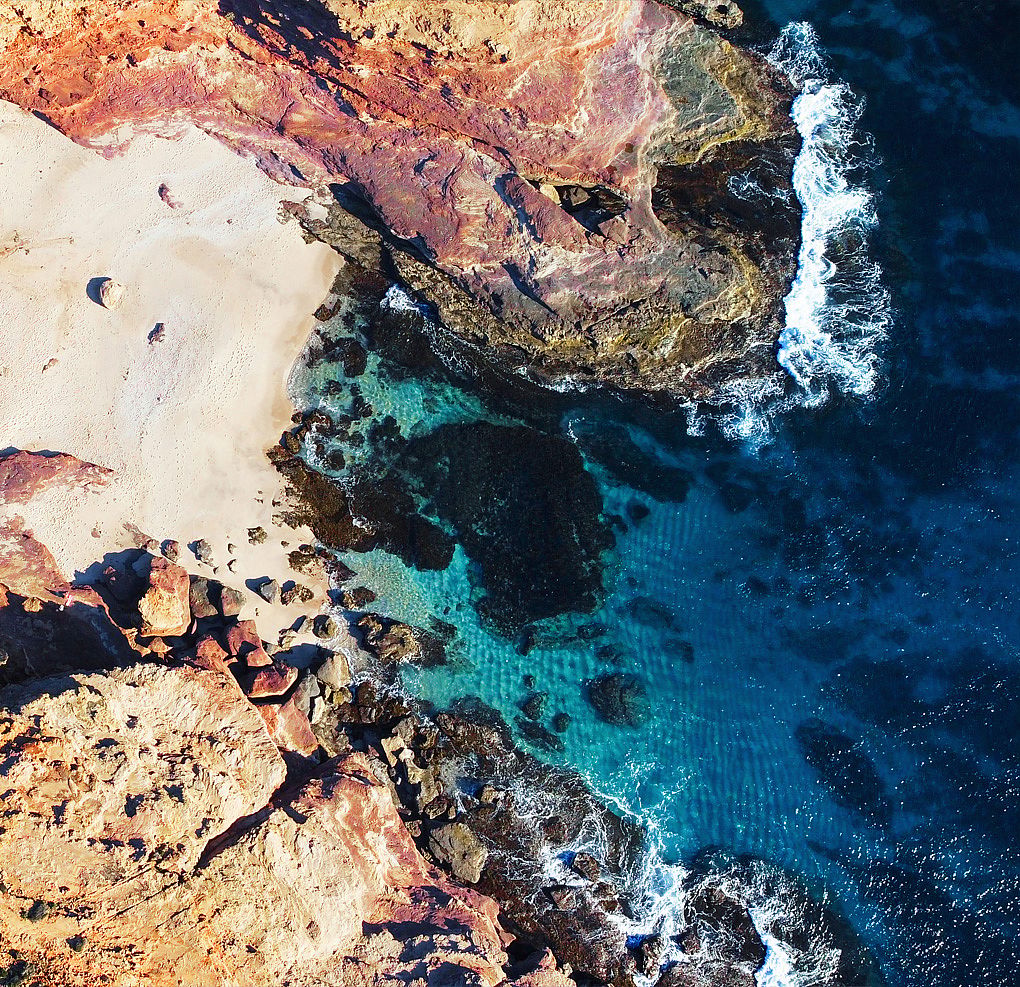 Luchtfoto van een rotsachtige kustlijn met bleek zand, roestkleurige kliffen en helderblauwe zee.