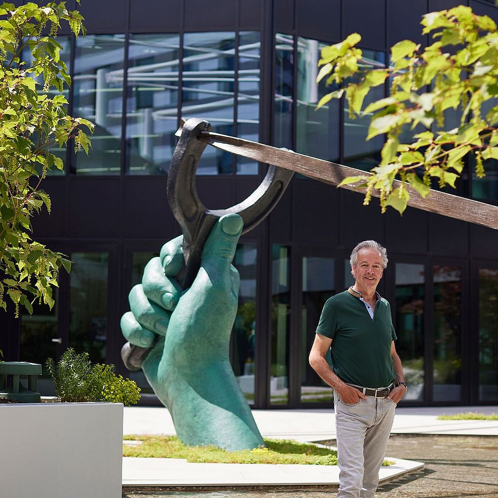 A man stands beside a large green sculpture of a hand holding a slingshot in front of the modern glass façade of the AFAS Experience Center.