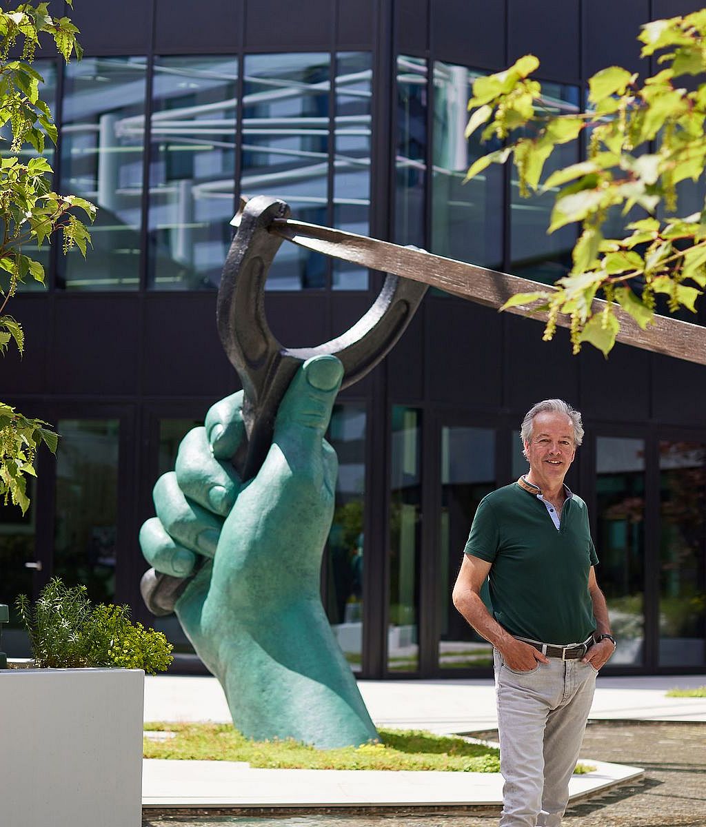 Een man staat naast een grote groene sculptuur van een hand met een katapult voor de moderne glazen gevel van het AFAS Experience Center.