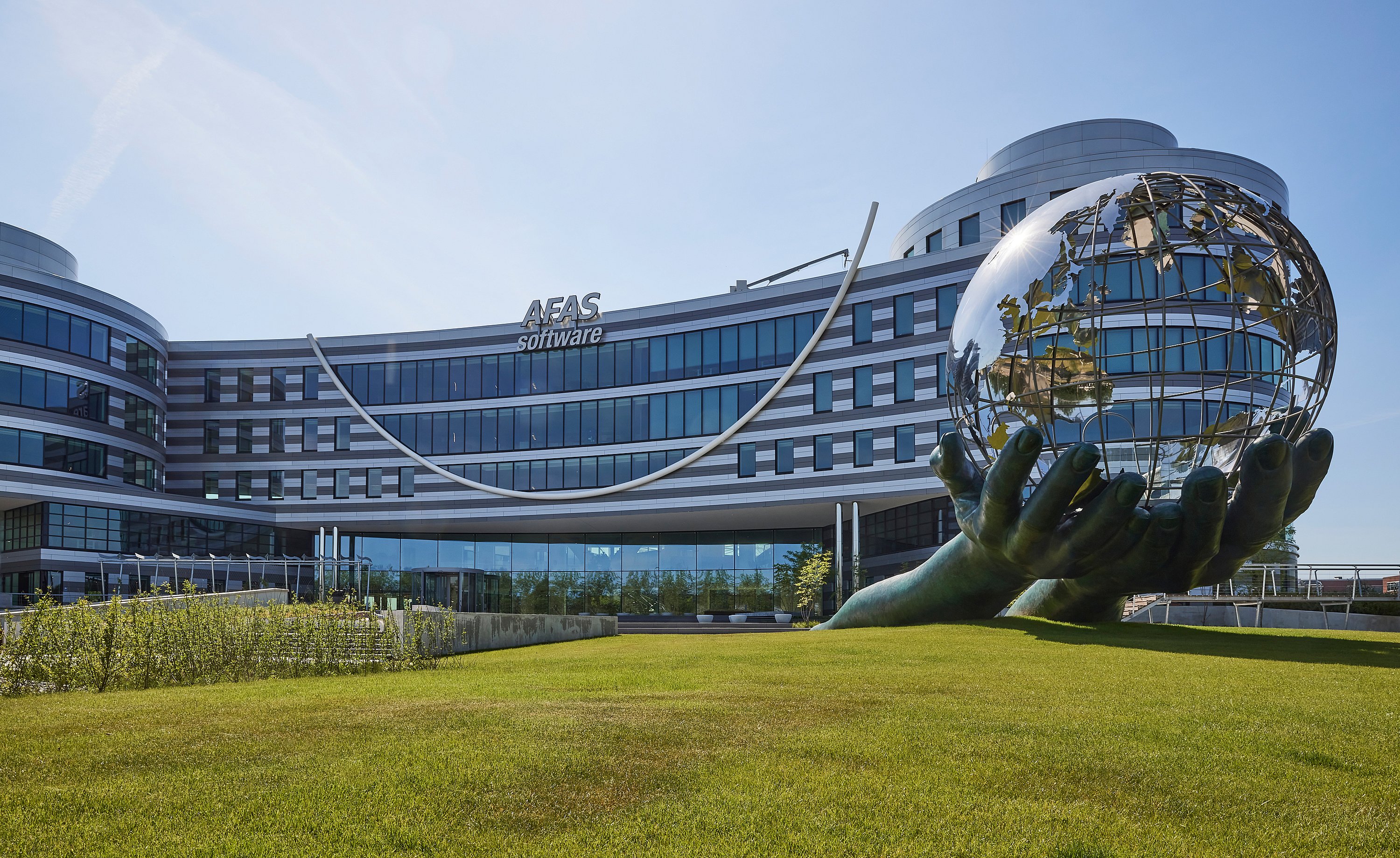 Large vue extérieure du bâtiment AFAS Software avec sa façade en verre incurvée, sa grande pelouse et une sculpture monumentale de mains tenant un globe.