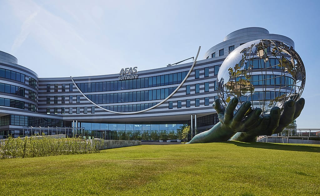 Wide exterior view of the AFAS Software building with its curved glass façade, large lawn and a monumental sculpture of hands holding a globe.