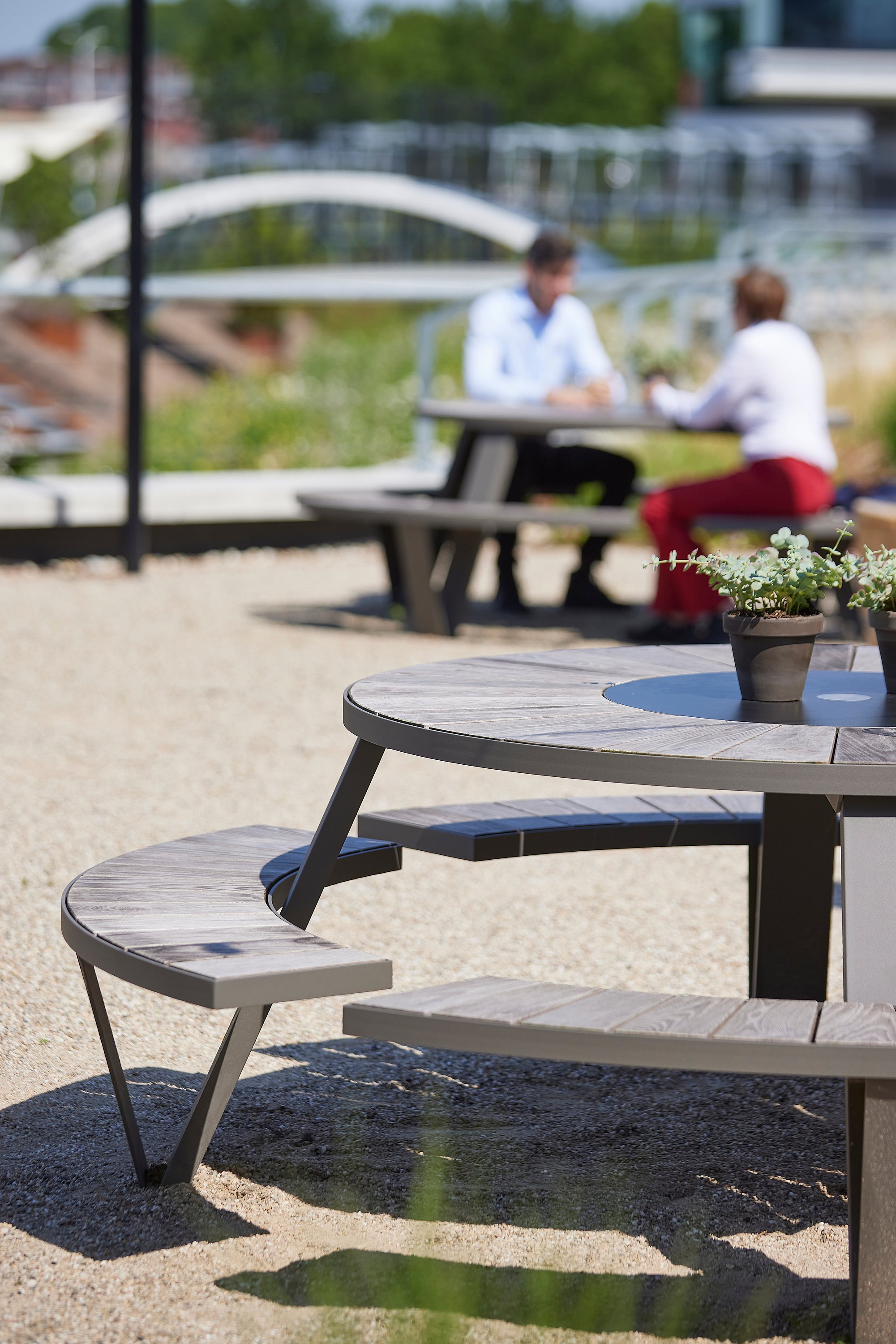 A grey Pantagruel picnic table stands on gravel in the foreground, while two people meet at another outdoor table near a bridge in the background.