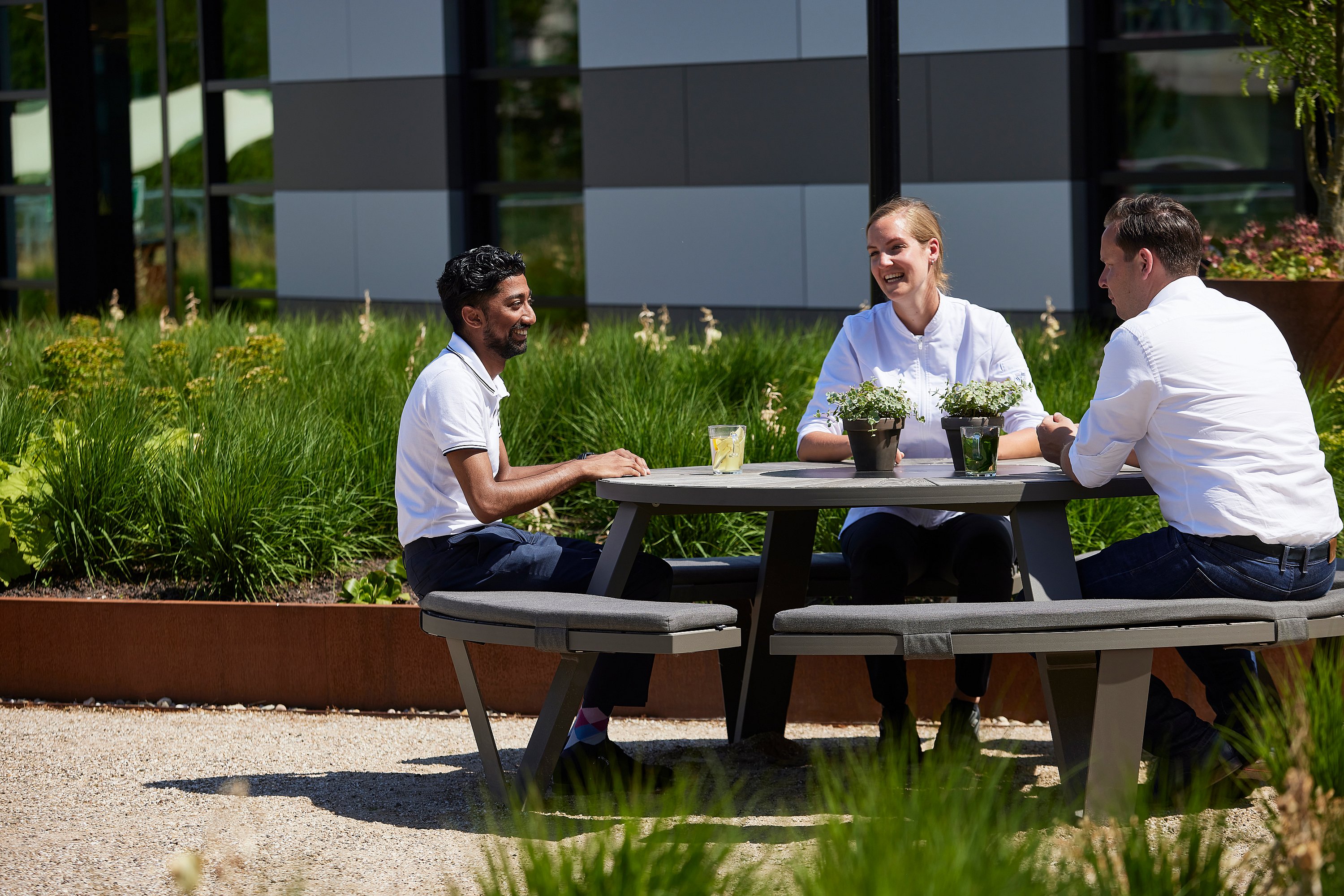 Trois collègues sont assis ensemble à une table de pique-nique grise Pantagruel dans un jardin paysager avec des herbes et le bâtiment AFAS derrière.