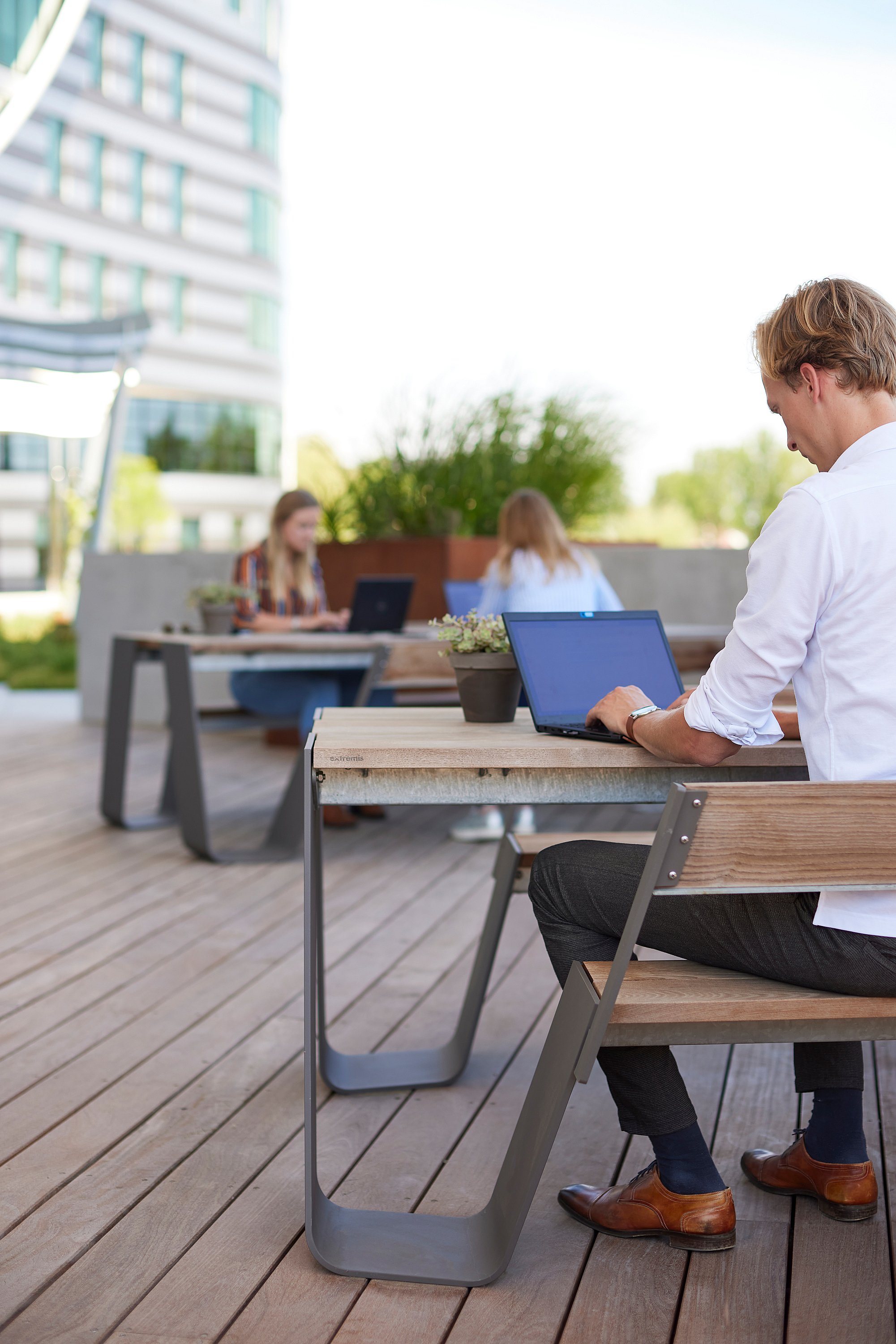 Un homme travaille sur un ordinateur portable à une table en bois sur une terrasse ensoleillée, avec deux collègues utilisant une autre table en arrière-plan.