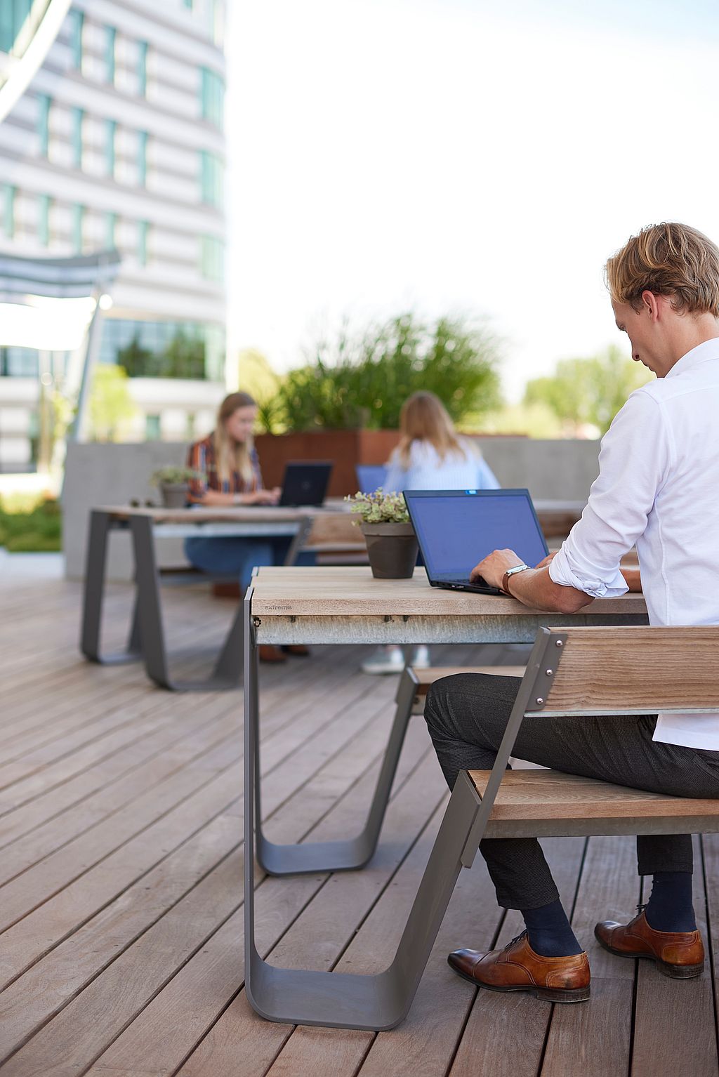 A man works on a laptop at a wood-topped Hopper table on a sunlit terrace, with two coworkers using another table in the background